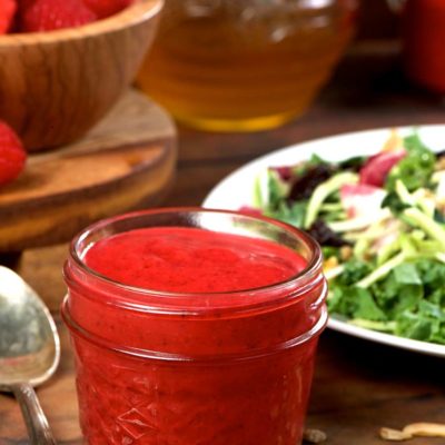 A small glass jar filled with raspberry vinaigrette with a spoon and side salad.