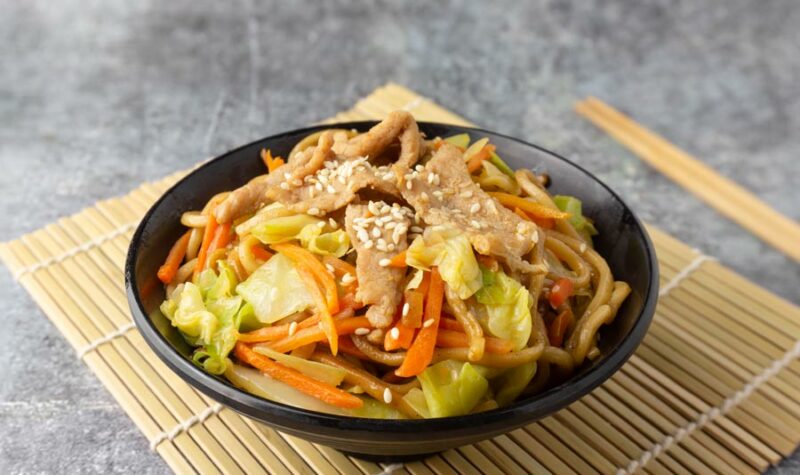 A black bowl filled with noodles and asian veggies, and pork.